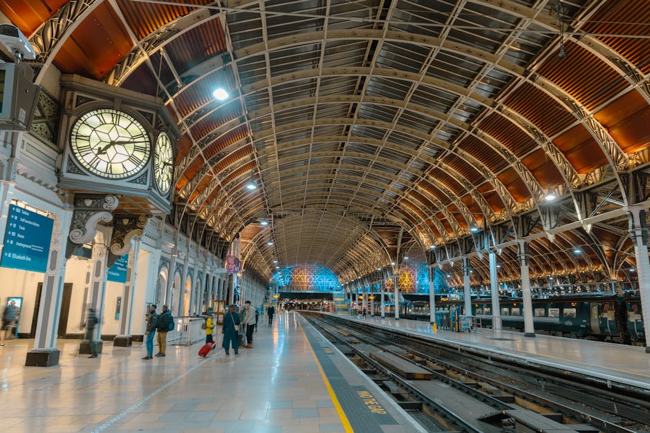 Inside Paddington Station's spacious platform area with a high, arched glass and metal roof illuminated by overhead lighting. On the left side, there are decorative clocks mounted on ornate white colu