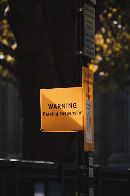 Close-up of a rectangular white metal sign affixed with four screws, displaying the message 'NO PARKING DAY OR NIGHT' in bold black capital letters, mounted on a grey wooden door or wall, likely at a